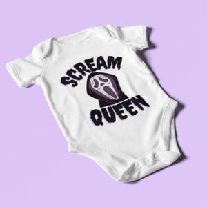 Puede incluir: Body de beb&eacute; blanco con el texto "SCREAM QUEEN" en negro y morado. El dise&ntilde;o presenta una cara de fantasma en una capucha negra. El body tiene mangas cortas y corchetes en la parte inferior.