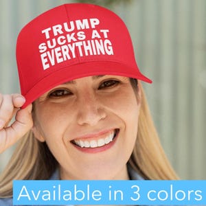 May include: A red baseball cap with the text "TRUMP SUCKS AT EVERYTHING" in white. The cap is being held up by a person. The image also includes the text "Available in 3 colors".