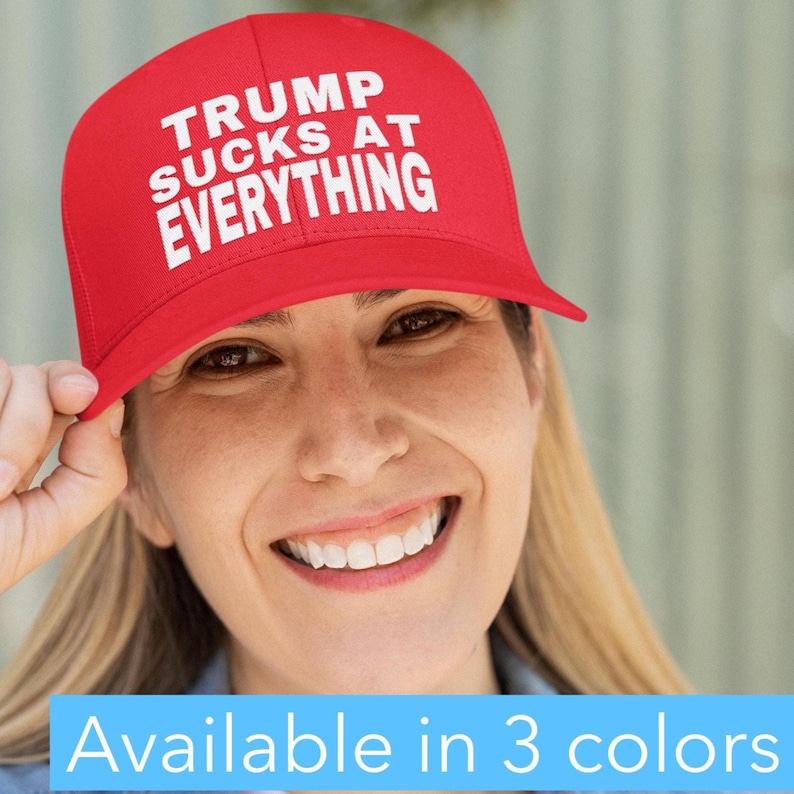 May include: A red baseball cap with the text "TRUMP SUCKS AT EVERYTHING" in white. The cap is being held up by a person. The image also includes the text "Available in 3 colors".