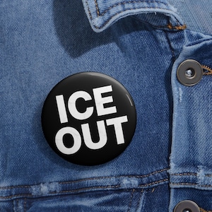 May include: A black button with the white text "ICE OUT" is pinned to a blue denim jacket. The button is round and has a glossy finish. The denim jacket has visible stitching and metal buttons.
