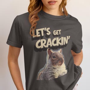 May include: Dark grey t-shirt with a squirrel graphic and the text "LET'S GET CRACKIN'". The squirrel is depicted in grey and white, with a vintage font. The shirt has short sleeves and a crew neck.