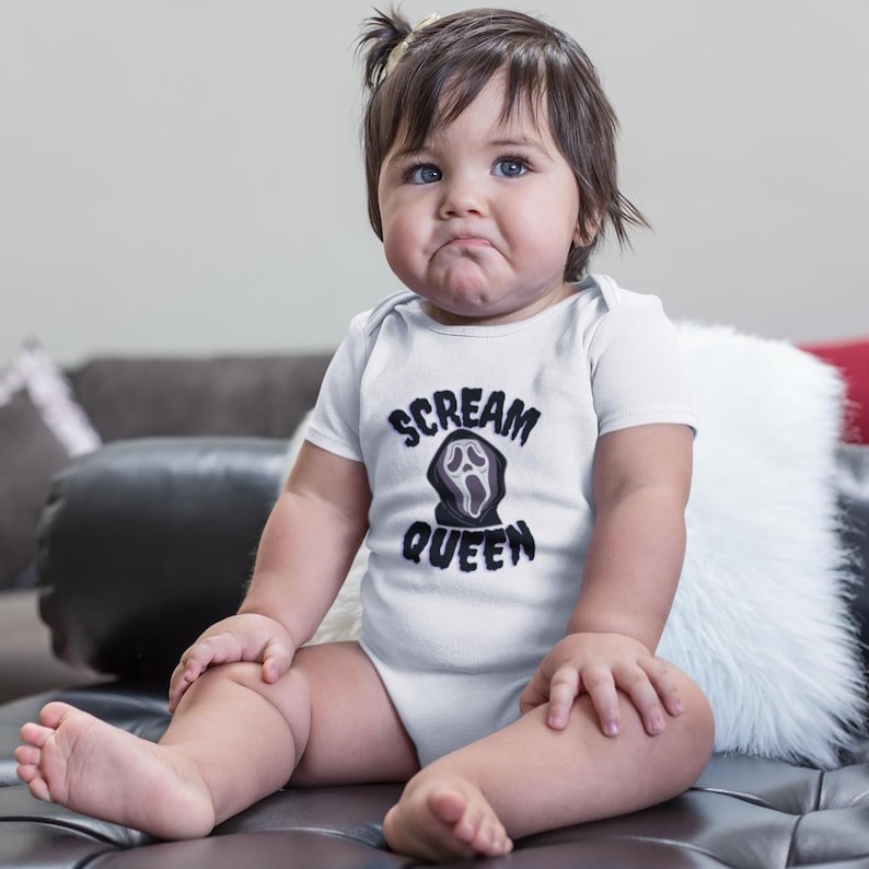 Puede incluir: Un body blanco para beb&eacute; con el texto "SCREAM QUEEN" en negro, con un dise&ntilde;o de cara de fantasma. El beb&eacute; est&aacute; sentado en un sof&aacute; de cuero negro con un coj&iacute;n blanco y esponjoso.