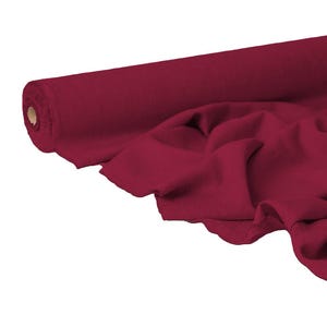 Può includere: Un rotolo di tessuto rosso scuro, parzialmente srotolato per rivelare la morbida consistenza e il drappeggio del materiale. Il tessuto è di colore uniforme, con una leggera lucentezza, e sembra essere di peso medio. Il rotolo è su sfondo bianco.