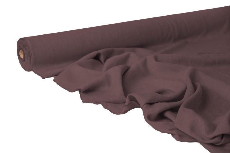 May include: A roll of dark mauve fabric with a soft, flowing drape. The fabric is partially unrolled, revealing the texture and folds of the material. The fabric appears to be a medium weight, suitable for various sewing projects.