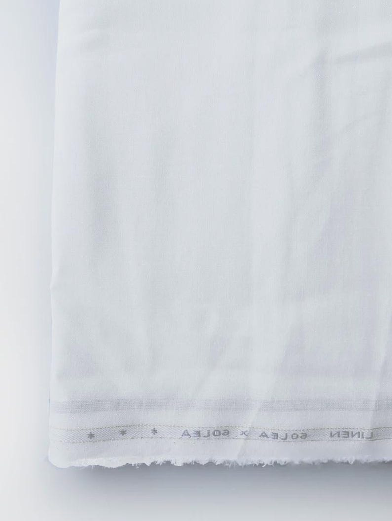 Peut inclure: Un tissu blanc pli&eacute; avec un bord brut. Le tissu a une texture subtile et une bordure avec du texte et des motifs en forme d'&eacute;toiles. Le tissu semble &ecirc;tre du lin ou une mati&egrave;re similaire, adapt&eacute; aux v&ecirc;tements ou &agrave; la d&eacute;coration int&eacute;rieure.
