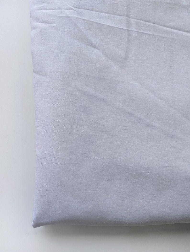 May include: A folded, light-colored fabric, possibly linen or cotton, with visible texture and subtle shadows. The fabric appears to be a solid, pale color, possibly white or a very light gray, and is neatly folded.