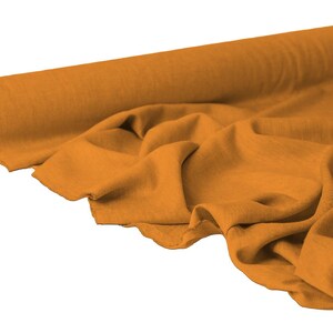 May include: A roll of mustard-colored fabric with a soft, flowing drape. The fabric is partially unrolled, revealing the texture and folds of the material. The background is plain white.
