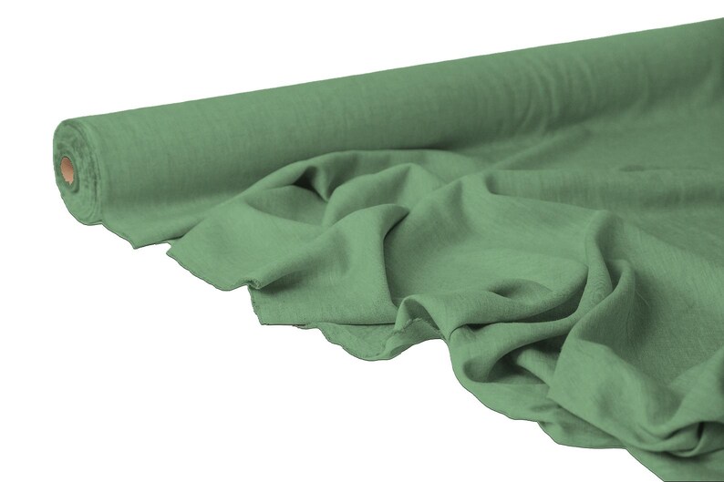 May include: A roll of sage green fabric with a soft, flowing texture. The fabric is partially unrolled, revealing its drape and the texture of the material. The roll has a cardboard core.