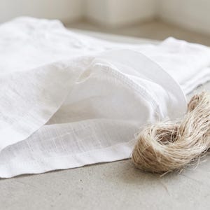 May include: A folded white linen shirt with a visible woven texture and a round neckline, accompanied by a small, coiled bundle of natural tan jute twine resting beside the fabric on a neutral, light-coloured surface.