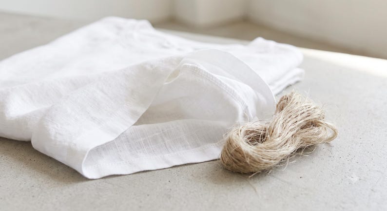 May include: A folded white linen shirt with a visible woven texture and a round neckline, accompanied by a small, coiled bundle of natural tan jute twine resting beside the fabric on a neutral, light-coloured surface.