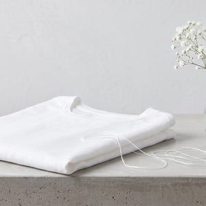 May include: A folded plain white t-shirt with a crew neckline. A thin white string lies across the fabric. Beside the shirt is a small clear glass bottle containing a sprig of white baby's breath flowers and a smooth grey stone.