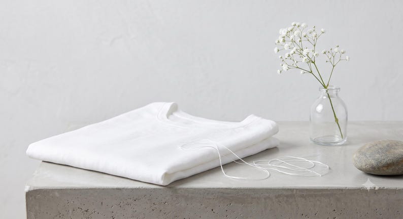 May include: A folded plain white t-shirt with a crew neckline. A thin white string lies across the fabric. Beside the shirt is a small clear glass bottle containing a sprig of white baby's breath flowers and a smooth grey stone.