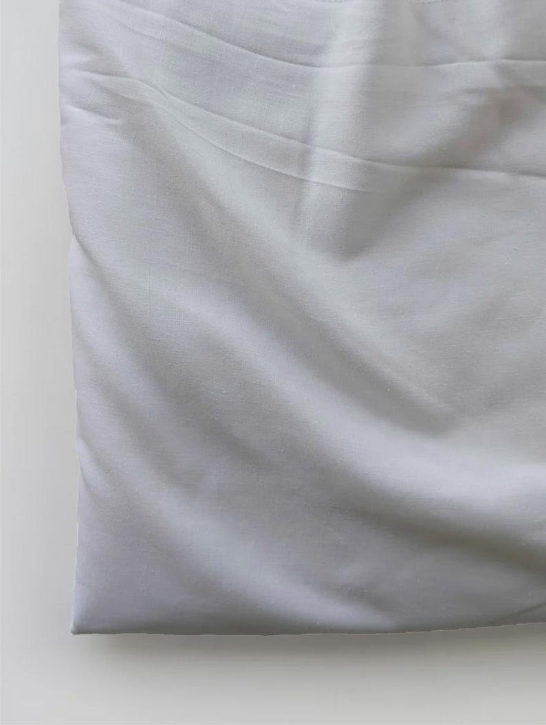 May include: A folded, white fabric with soft folds and subtle shadows. The material appears to be smooth and possibly cotton or linen, suggesting bedding or clothing.