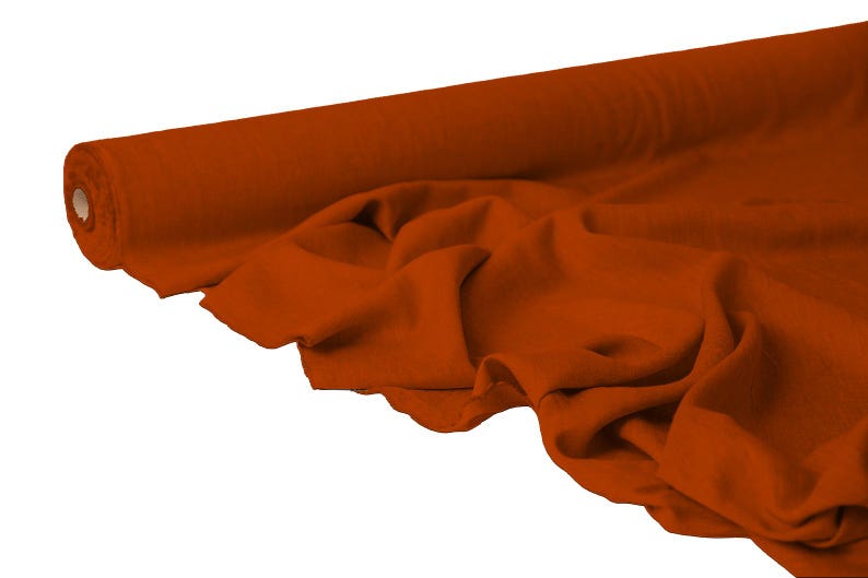 May include: A roll of rust-colored fabric, partially unrolled to reveal the soft texture. The fabric is draped in gentle folds, showcasing its flexibility and potential for various sewing projects. The color is a warm, earthy tone.