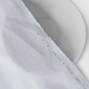 Peut inclure: Gros plan sur un tissu blanc avec un bord brut, drap&eacute; sur une assiette blanche. Le tissu pr&eacute;sente une texture subtile et une bordure imprim&eacute;e avec le texte "INTIMATE LINEN X GOLEA". L'image est bien &eacute;clair&eacute;e, mettant en valeur les d&eacute;tails du tissu.
