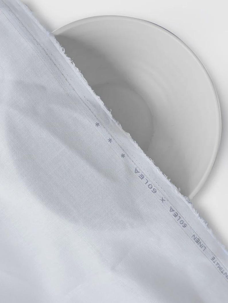 Peut inclure: Gros plan sur un tissu blanc avec un bord brut, drap&eacute; sur une assiette blanche. Le tissu pr&eacute;sente une texture subtile et une bordure imprim&eacute;e avec le texte "INTIMATE LINEN X GOLEA". L'image est bien &eacute;clair&eacute;e, mettant en valeur les d&eacute;tails du tissu.