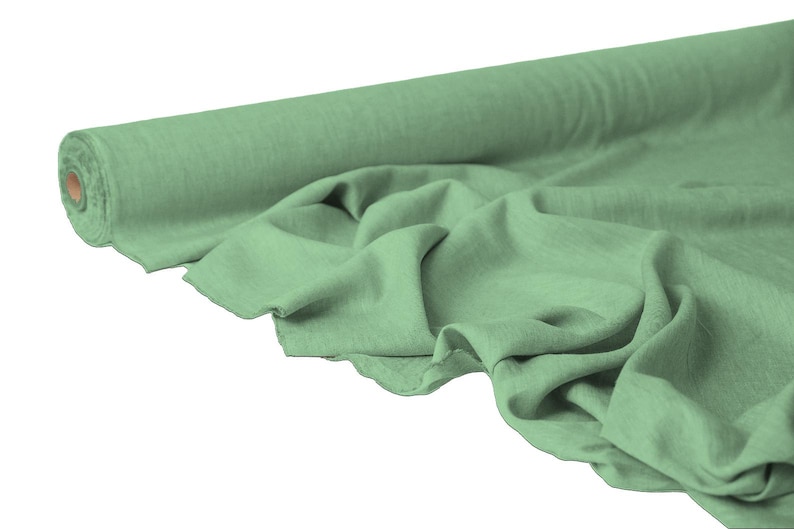May include: A roll of sage green fabric with a soft, textured appearance. The fabric is partially unrolled, displaying its draped folds. The material appears to be lightweight and suitable for various sewing projects.