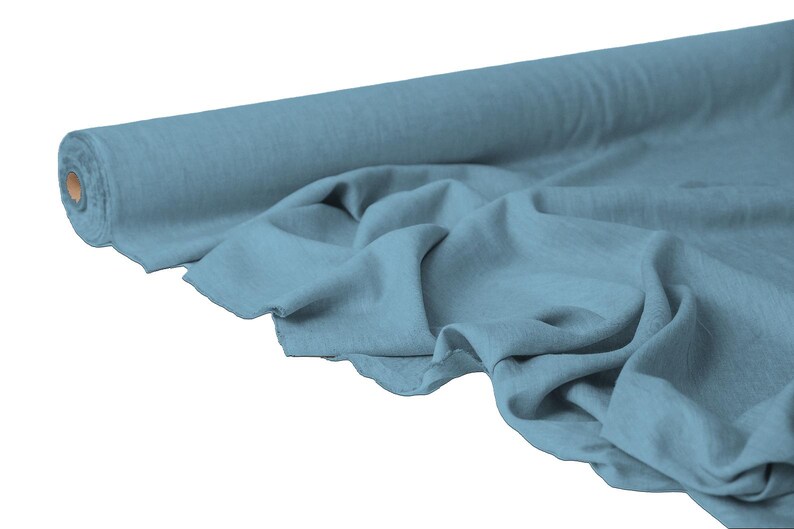 May include: A roll of light blue fabric with a soft, textured appearance. The fabric is partially unrolled, displaying its draped folds. The core of the roll is visible, showing a brown cardboard center. The fabric appears to be a lightweight material.