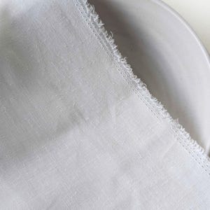 May include: A close-up of a white linen fabric with a frayed edge, partially covering a light gray ceramic plate. The fabric has a textured appearance and a subtle weave pattern, suggesting a natural fiber.