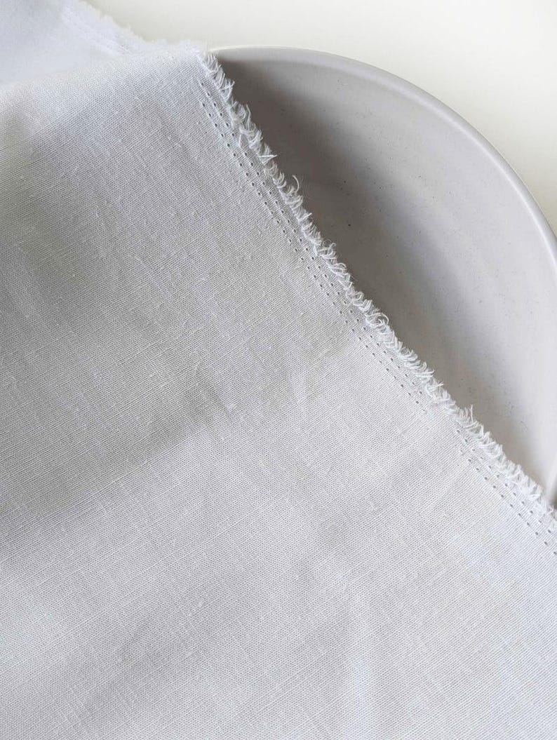 May include: A close-up of a white linen fabric with a frayed edge, partially covering a light gray ceramic plate. The fabric has a textured appearance and a subtle weave pattern, suggesting a natural fiber.