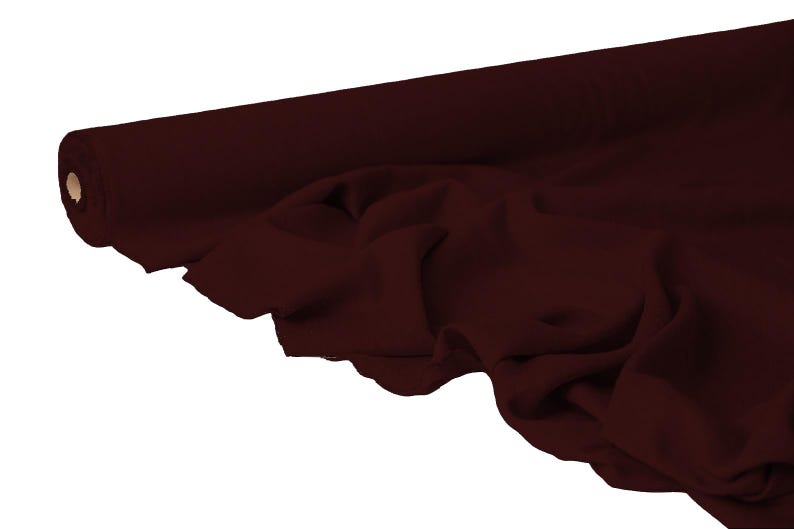 May include: A roll of burgundy fabric, partially unrolled to reveal the texture and drape of the material. The fabric is a deep, rich shade of red-brown, with soft folds and a smooth appearance. The roll is positioned on a white background.