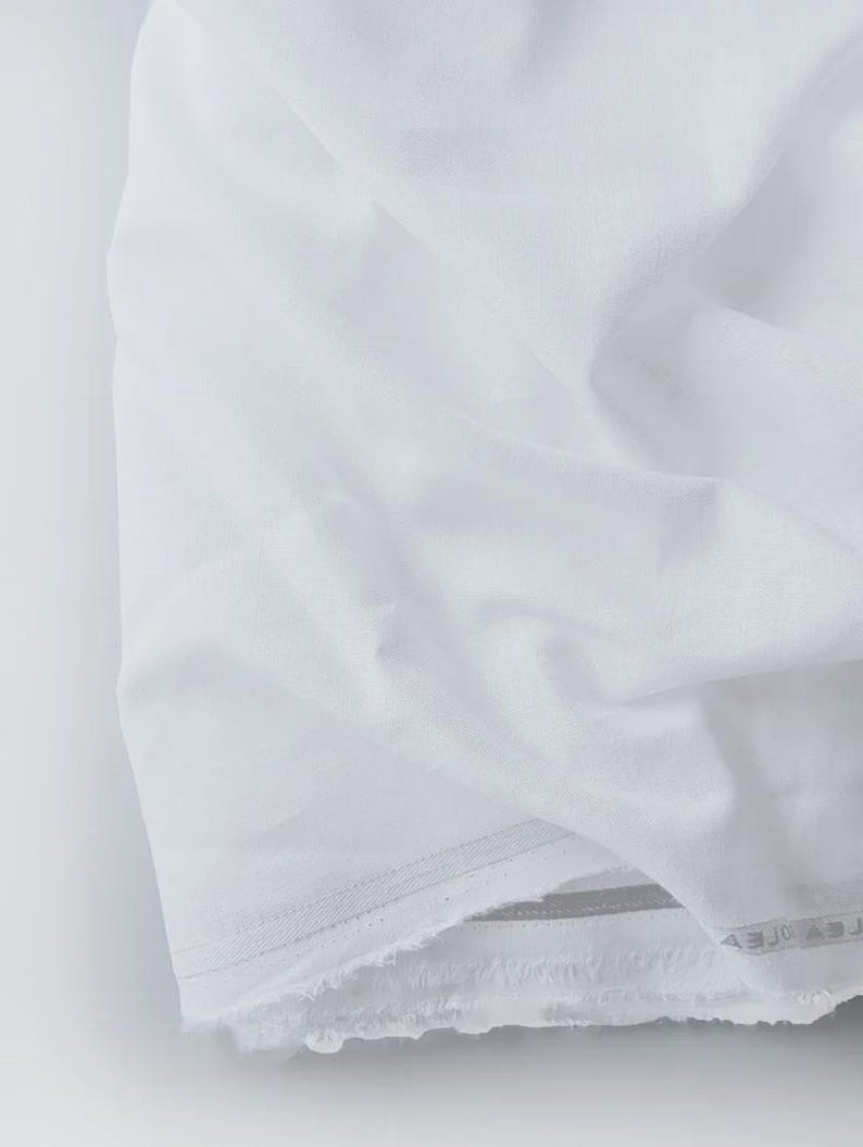 Peut inclure: Gros plan sur un tissu blanc pli&eacute;. Le tissu pr&eacute;sente une texture douce avec des plis et des froissements visibles. Les bords sont l&eacute;g&egrave;rement effiloch&eacute;s, pour un aspect simple et &eacute;pur&eacute;. Le tissu semble &ecirc;tre du coton ou du lin.