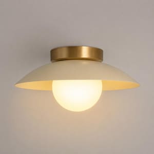 May include: A flush mount ceiling light fixture featuring a circular brass canopy and a wide, shallow, off-white metal shade. A spherical, frosted glass globe bulb is positioned centrally beneath the shade, emitting a soft, warm, diffused light.