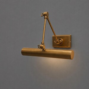 May include: A brass-toned wall-mounted picture light. The light features a cylindrical shade, adjustable arm, and a rectangular wall plate. The design is minimalist and industrial, suitable for illuminating artwork or decor.