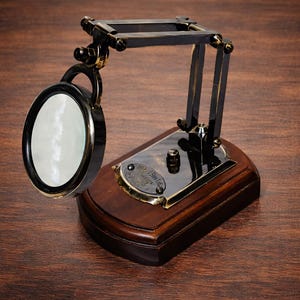 May include: A vintage-style magnifying glass with a round lens, black frame, and a wooden base. The magnifying glass is attached to a black and gold metal arm. The base has a brass plate with text.