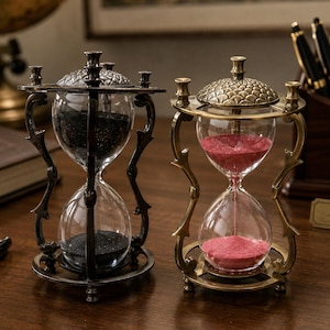 May include: Two hourglasses with ornate metal frames and textured dome tops. The left hourglass contains black sand, while the right hourglass contains pink sand. Both feature clear glass bulbs held by curved, decorative metal supports on circular bases.