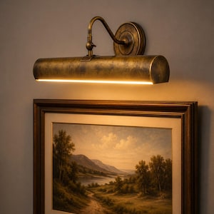 May include: A wall-mounted picture light with an antique brass finish and a cylindrical shade. The fixture features an adjustable arm and emits a warm, downward-facing glow, illuminating a framed landscape painting of a river valley below.
