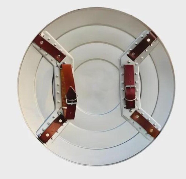 May include: A silver-colored, round shield with concentric circles. It features brown leather straps and silver metal brackets secured with rivets. The shield has a classic, vintage design.