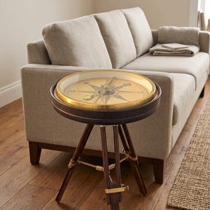 May include: A round side table featuring a dark wood frame and a glass top that encases a large, gold-toned nautical compass dial. The table is supported by three dark wood legs connected by brass hardware, resembling a vintage tripod stand.
