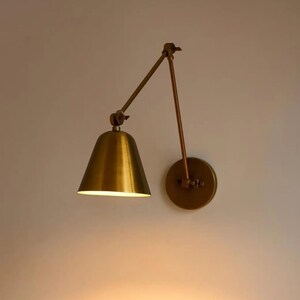 May include: A wall-mounted lamp featuring a metallic brass finish with a conical shade and an adjustable swing arm. The fixture includes a circular wall plate and articulated joints that allow for positioning the light source as needed.