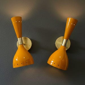 May include: Two mid-century modern wall sconces. The lamps have a bright yellow, conical shade with small holes and a brass-colored base. The lamps are mounted on a dark gray wall.