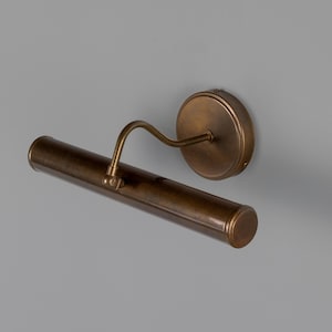 May include: A wall-mounted picture light with a cylindrical, antique brass-finished metal shade. The fixture features a circular wall plate and a curved, adjustable arm that connects to the center of the horizontal tube-shaped light housing.