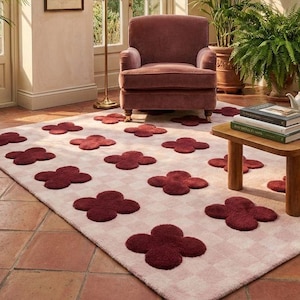 Pode incluir: Uma sala de estar com um tapete estampado com desenhos de trevos bordô sobre um fundo xadrez rosa claro. Uma poltrona bordô está no fundo, com uma pequena mesa de madeira e plantas. O ambiente é acolhedor e convidativo.
