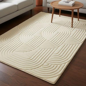 Puede incluir: Alfombra de color crema con un patrón geométrico de líneas curvas. La alfombra está colocada sobre un suelo de madera oscura, con dos sofás grises y una mesa de centro de madera en el fondo. También se ve una planta en maceta.
