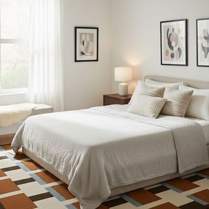 May include: A bedroom scene featuring a bed with a light gray blanket and pillows. A geometric patterned rug in brown, blue, and cream tones is on the floor. Artwork hangs on the white walls, and a lamp sits on a wooden nightstand.
