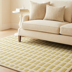 May include: A cream-coloured sofa with two patterned cushions rests on a rug featuring a repeating rectangular design in light green. The rug is on a wooden floor, and a small side table is visible.