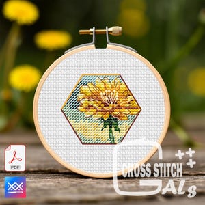 Puede incluir: Un bastidor de bordado de punto de cruz con una flor de diente de león amarilla en un marco hexagonal. El diseño está sobre una tela blanca, sujeta dentro de un aro de madera. La imagen incluye el texto "CROSS STITCH GALS".