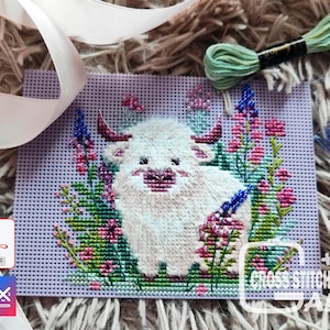 Peut inclure: Un projet de point de croix représentant un yak blanc entouré de fleurs roses, violettes et bleues. Le yak a des cornes roses et un nez rose. Le projet est sur un fond lavande. L'image comprend du fil à broder, des perles et le texte "CROSS STITCH ALS".