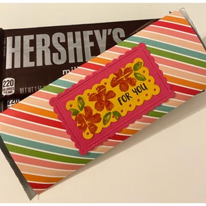May include: A Hershey's milk chocolate bar partially wrapped in a colorful striped paper sleeve. The sleeve has a pink scalloped label with the words "FOR YOU" and floral designs.