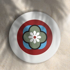 May include: A white, round decorative plate with a floral design. The design features a central green and white flower shape, surrounded by blue, red, and black accents. The plate is set against a sandy background with shadows.