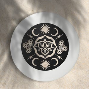 May include: A silver-colored, round decorative plate with a black center featuring a symmetrical design of moons, stars, suns, and floral elements. The design is in a cream color, creating a contrast against the black background. The plate is set against a sandy background.