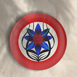 May include: A red, round plate with a floral design. The center of the plate features a blue and red flower with black accents on a white background. The plate is likely made of ceramic or a similar material.
