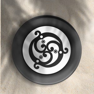 May include: A black and silver decorative object with a stylized, swirling design. The object is circular, with a black outer ring and a silver center featuring a black, symmetrical pattern. The design includes three interconnected, curved shapes.