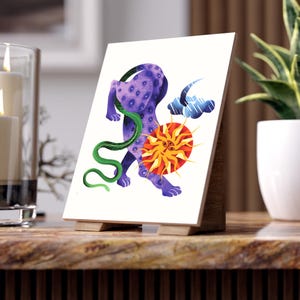 Puede incluir: Una impresión artística decorativa que presenta una criatura morada con una serpiente verde, un sol con ojos y una nube. La obra de arte se exhibe en un caballete de madera, ideal para la decoración del hogar.