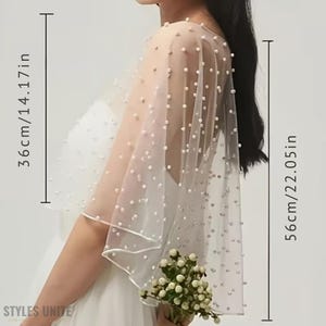 May include: A sheer, ivory-colored bridal cape, adorned with scattered pearl-like beads. The cape measures 14.17 inches (36cm) wide and 22.05 inches (56cm) long. The cape is worn over a white dress, and the bottom edge is trimmed with a delicate border.