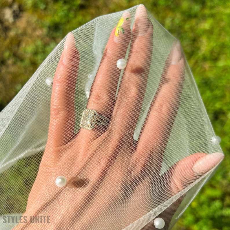 May include: A sheer white bridal veil adorned with scattered faux pearls. A hand wearing a diamond ring is visible through the veil. The text "STYLES UNITE" is at the bottom left corner.